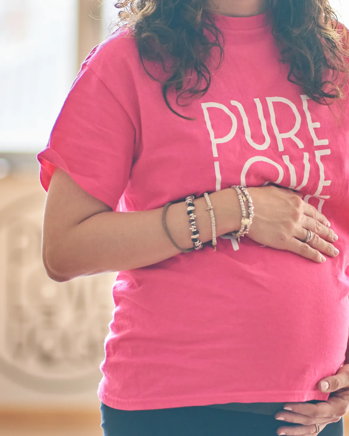 Una praticante incinta in maglietta Pure Love Yoga, le mani appoggiate sul pancione, con il logo della scuola sfocato sulla parete alle sue spalle