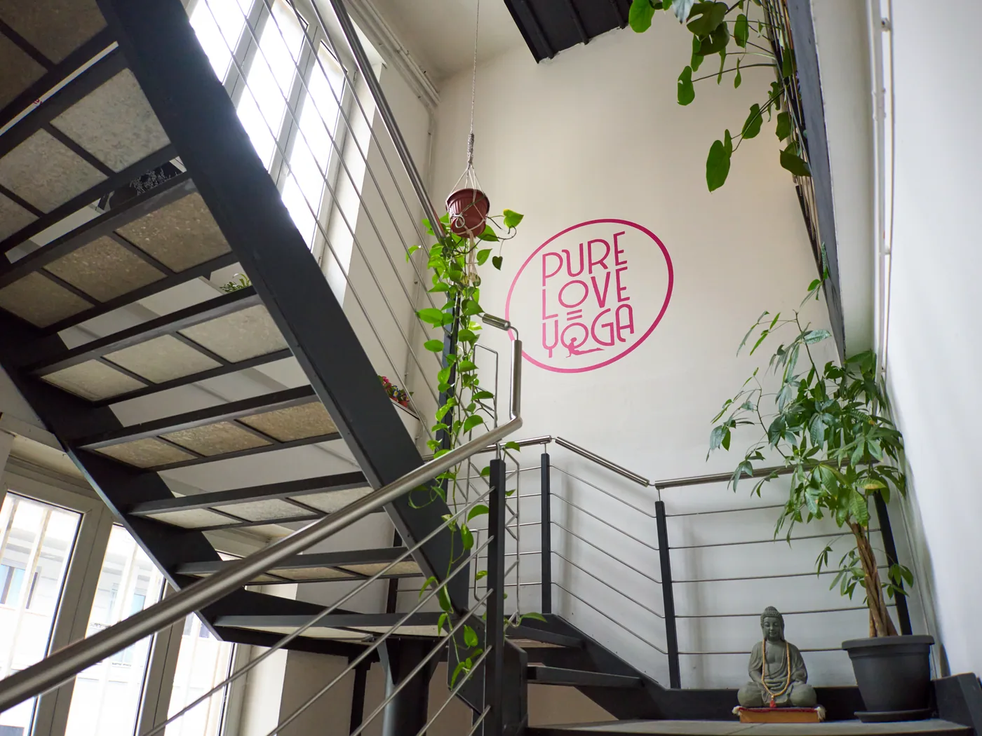La scala in acciaio che porta alle sale di Via Fattori, con il logo fucsia sulla parete, un piccolo Buddha di pietra e le piante