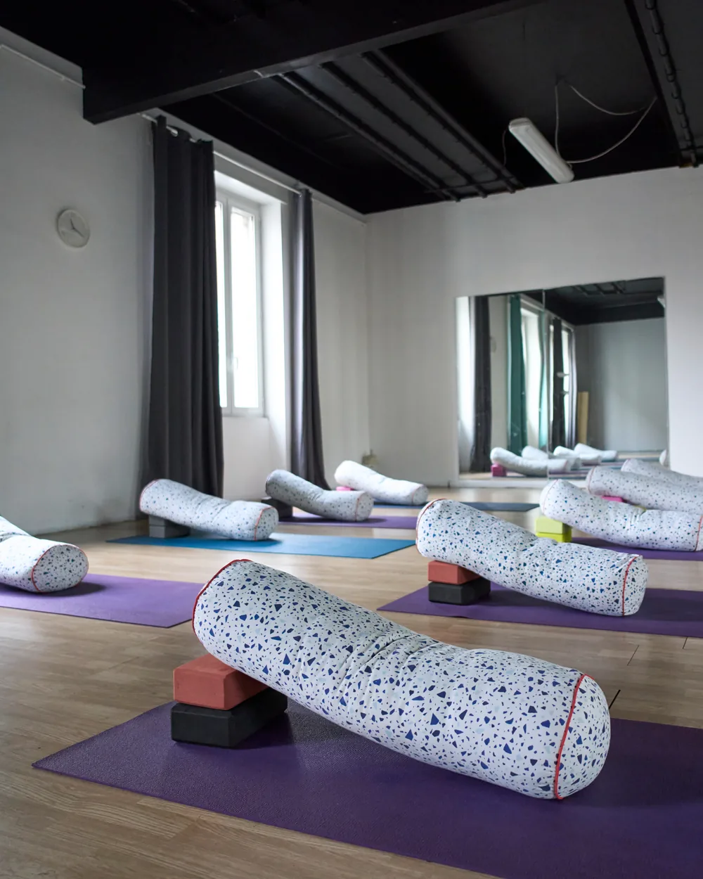 Sala 2 di Via Fattori allestita per una pratica lenta: bolster e mattoncini sui tappetini viola, tende verde acqua