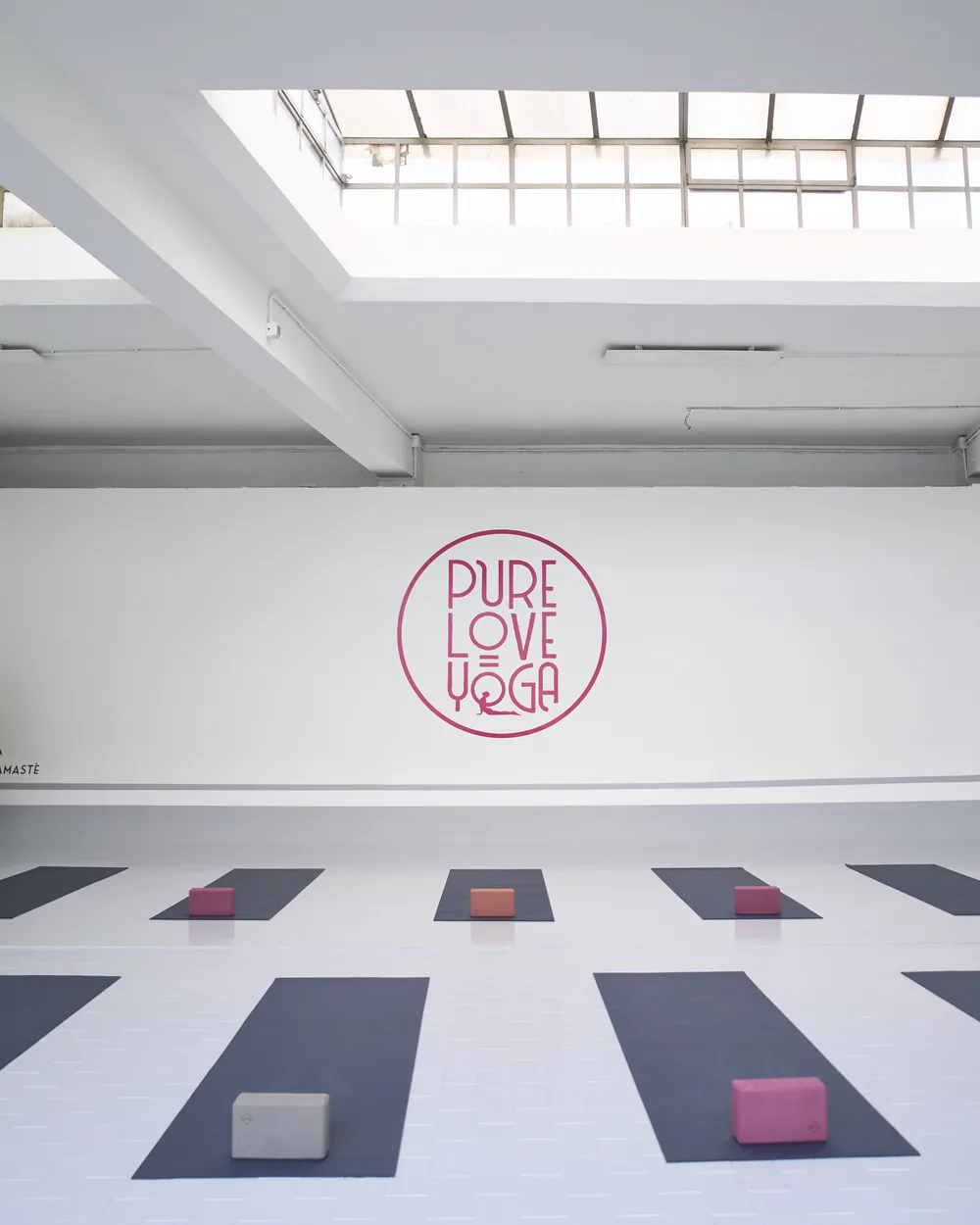 Lo Studio di Via Fattori: sala bianca con lucernari, logo Pure Love Yoga sulla parete e tappetini stesi a terra